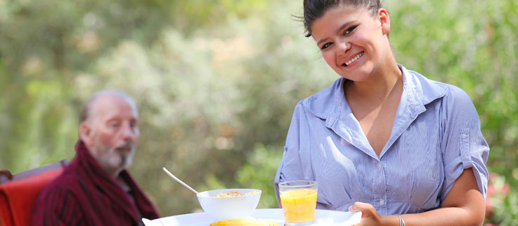 Female home care aide serves breakfast outside to elderly man sitting at table.