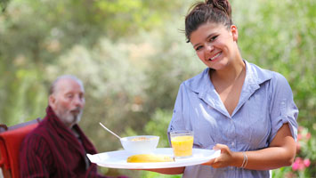 Female caregiver serves breakfast outside to elderly man sitting at table outside.