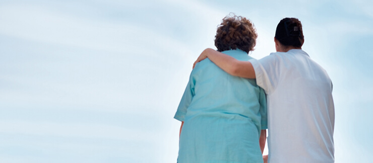 Female care aide puts arm around elderly woman as they both look up to the sky outside.