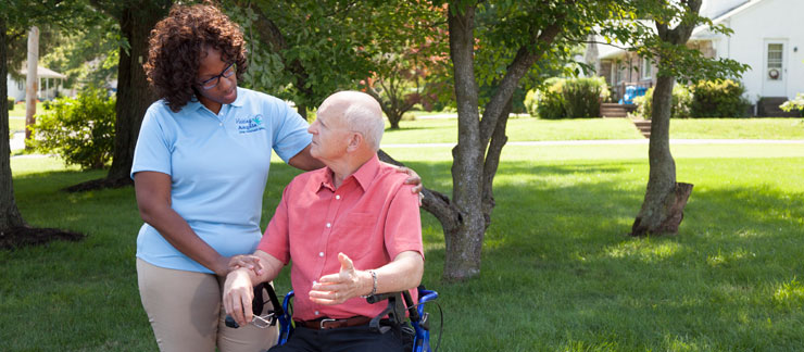 Female caregiver stands next to man with dementia sitting in wheelchair outside.