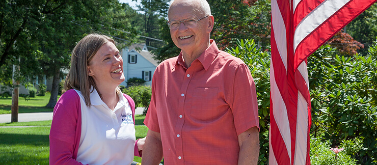 Female caregiver stands outside next to an elderly man by an American flag.