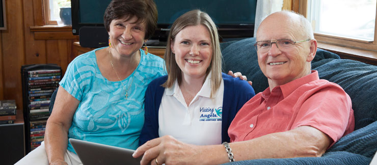 Female caregiver sits with senior couple on couch at home.