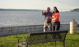 Elderly man holds the arm of a female home care worker as they walk along a path near the river.