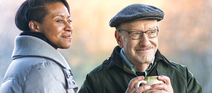 Female home care aide stands outside on a cold day with elderly man drinking hot coffee.