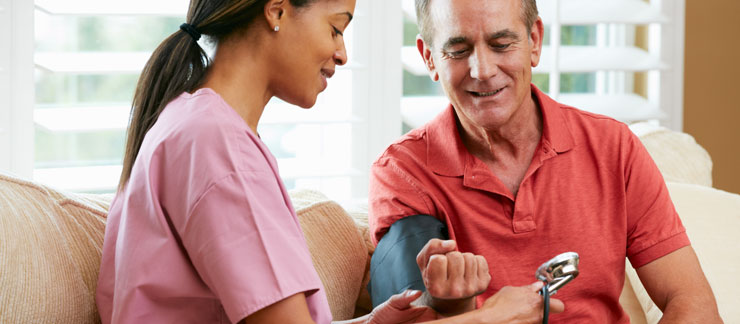 Female care aide checks senior man's blood pressure while sitting on couch.