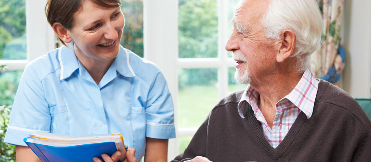 An in-home care provider stands next to a sitting elderly man as he drinks his coffee. The two are smiling at each other.