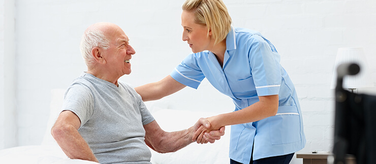 Elderly man in pain receives help trying to get up from bed by a female home care worker.