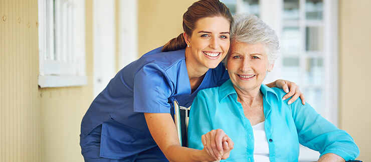 Female care worker embraces senior woman sitting outside of her home.