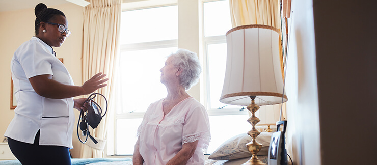 Female caregiver with blood pressure monitor tends to elderly woman sitting on bed.