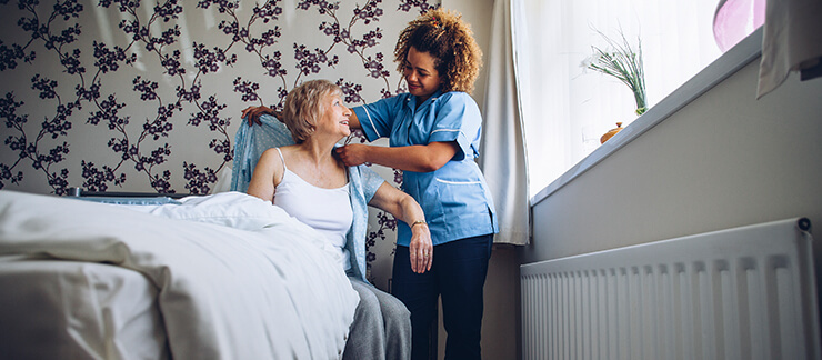 Female home care aide helps senior woman sitting in bed to put her shirt on.