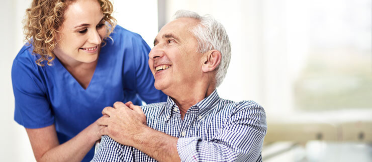 Female senior care worker holds the hand of older man sitting on a chair at home.