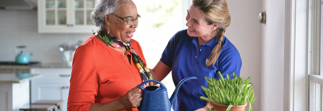 Mid-aged woman takes pride in her caregiver job by helping an elderly woman at home water a plant.
