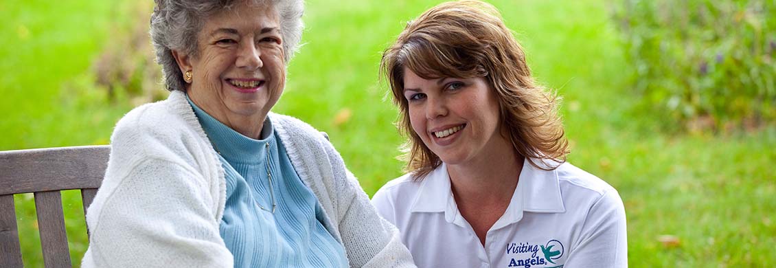 Experienced home care provider sits down outside with elderly woman during a nice afternoon.