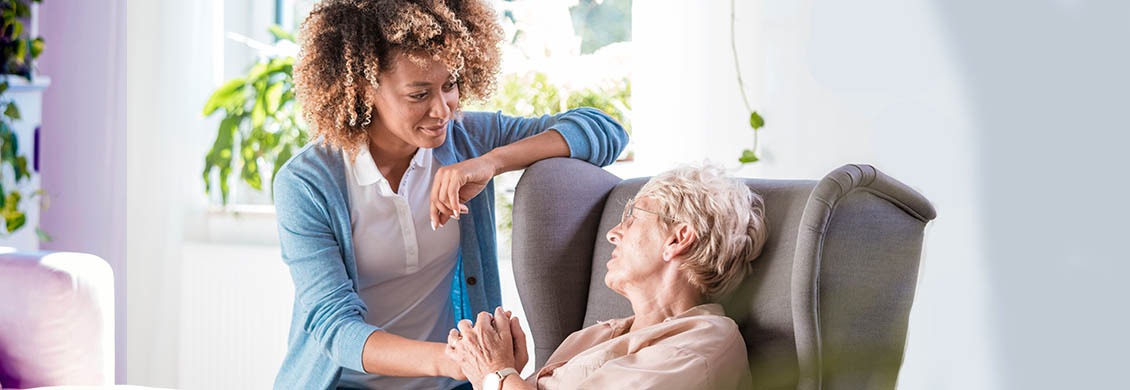 Senior woman sitting in chair and holding a female caregiver's hand.