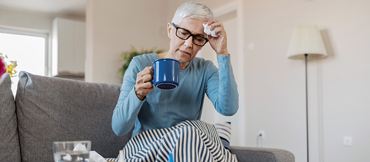 Senior woman with the flu is wrapped in a blanket and drinking hot tea while sitting on sofa at home.