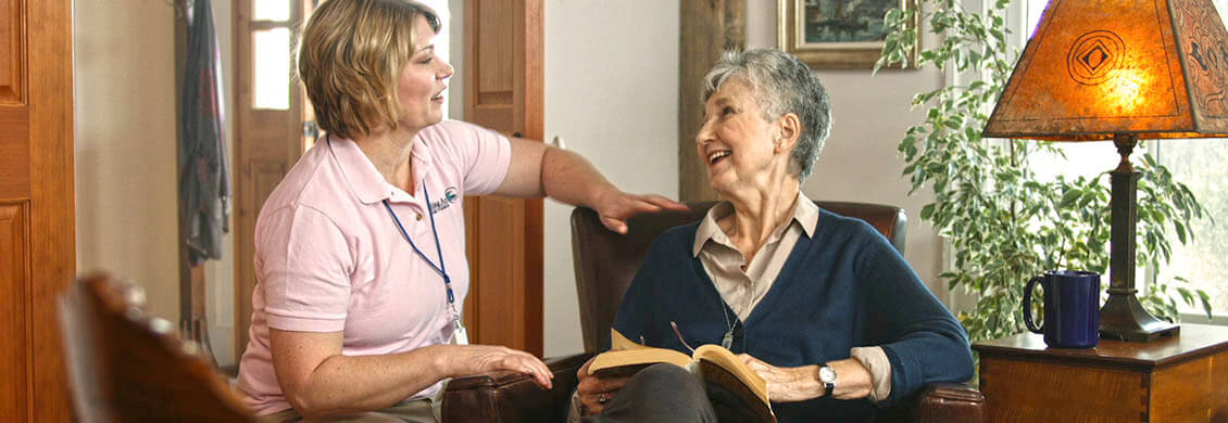 Home caregiver checking on an elderly woman happily reading a book on her couch.