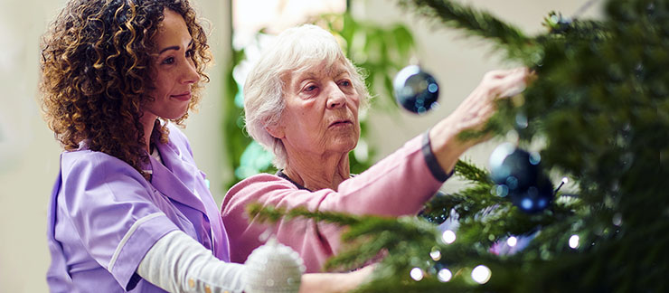 Female care worker helps elderly woman with decorating Christmas tree in her home during the holiday season.