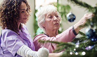 Female care worker helps elderly woman with decorating Christmas tree in her home during the holiday season.