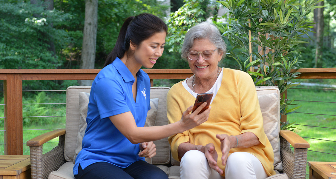 Female senior home care provider shows elderly woman a phone in the yard.