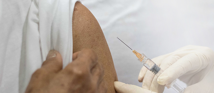 A closeup of an upper arm with rolled up sleeves is next to a gloved hand holding a needle 