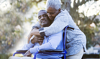 Senior woman hugs husband in wheelchair outside.