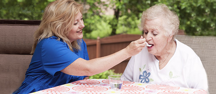 Female caregiver helps elderly woman with swallowing problem to drink water from a spoon.